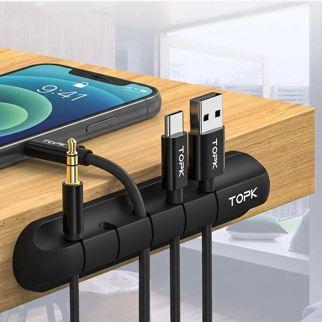 Black silicone cable clip holding cables on the edge of a wood desk, with a phone nearby
