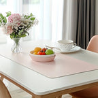 A pink desk mat on a white table with a bowl of fruit, a coffee cup, and a flower vase.