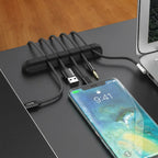 Black silicone cable clip on a desk edge holding USB cables, with a phone and laptop nearby