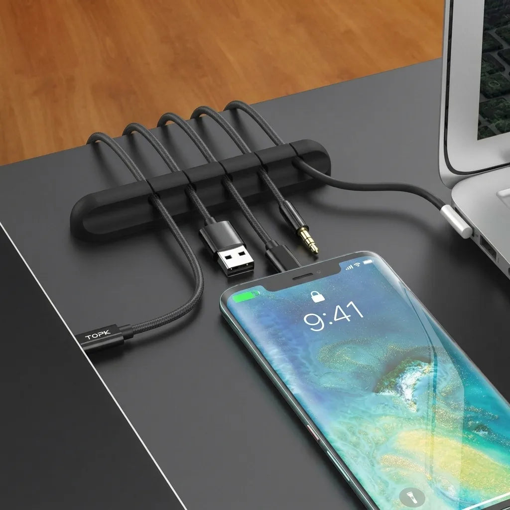Black silicone cable clip on a desk edge holding USB cables, with a phone and laptop nearby